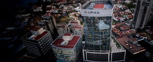 Vista aérea de un edificio corporativo moderno con el logotipo de UPAX en la parte superior, rodeado de otras construcciones y una zona urbana.